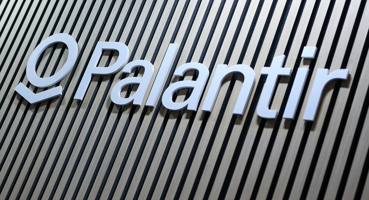 EPA/Shutterstock Close-up of the Palantir logo sign in white lettering mounted on a wall with vertical wooden slats in alternating light and dark tones.